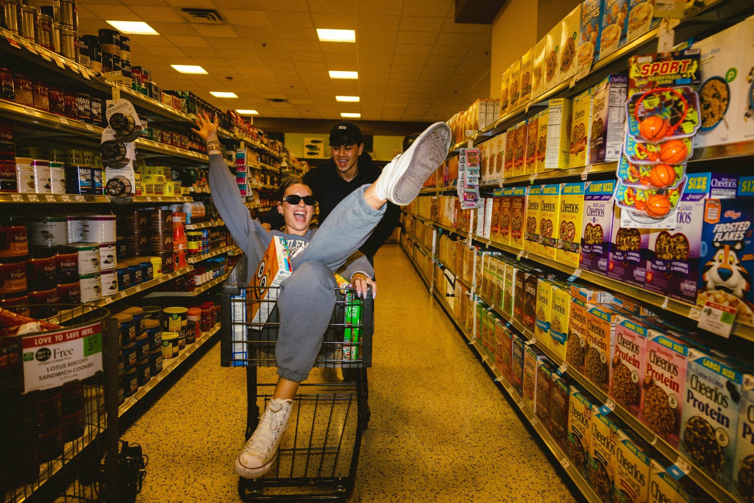 奥兰多活力超市里，两人推着购物车，一人在车外推着，另一人坐在车里，双腿俏皮地翘在空中。