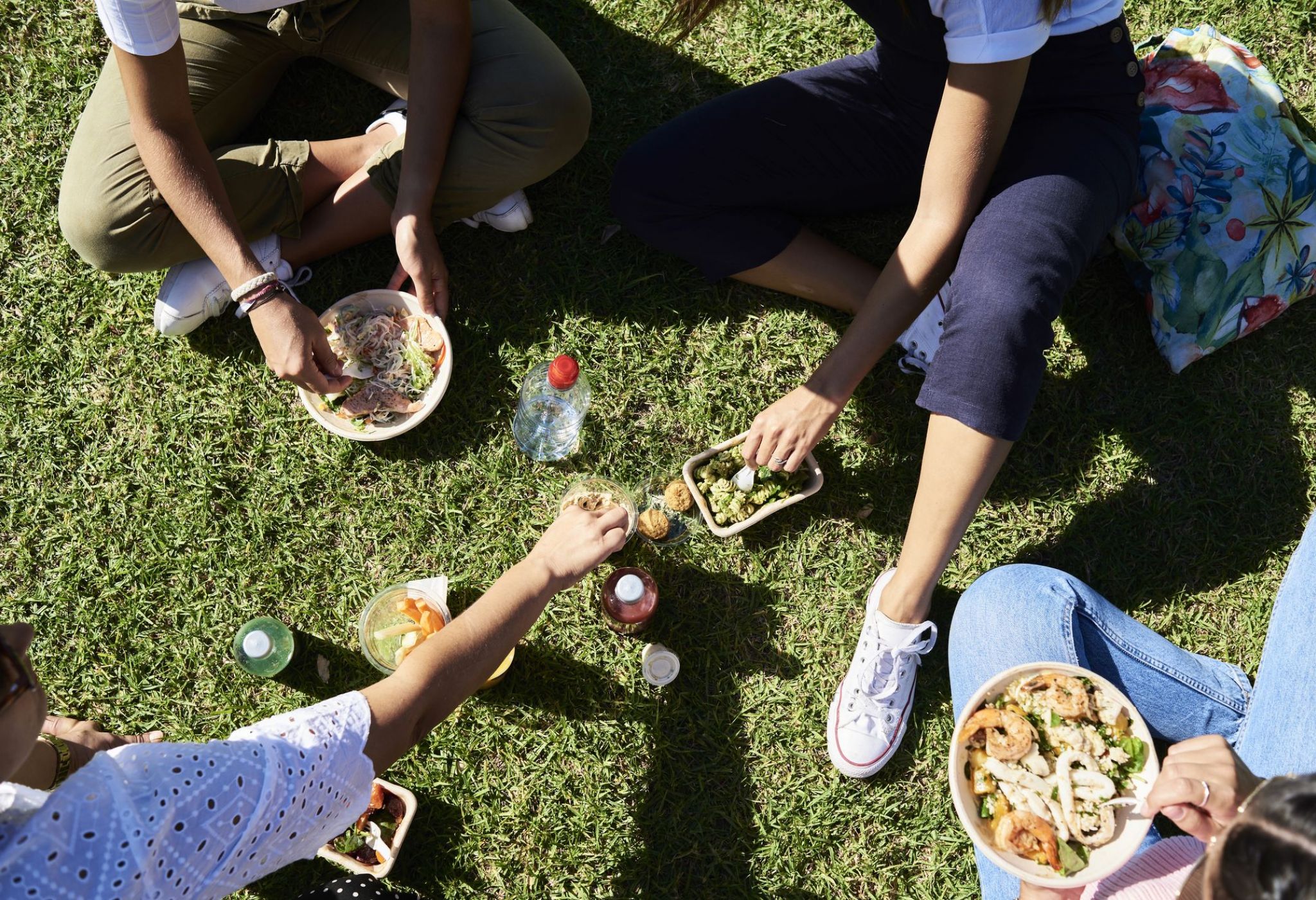 Overhead view of friends enjoying a casual picnic on the lawn with takeout bowls and drinks—capturing the sunny, social energy of life at VERVE Orlando.