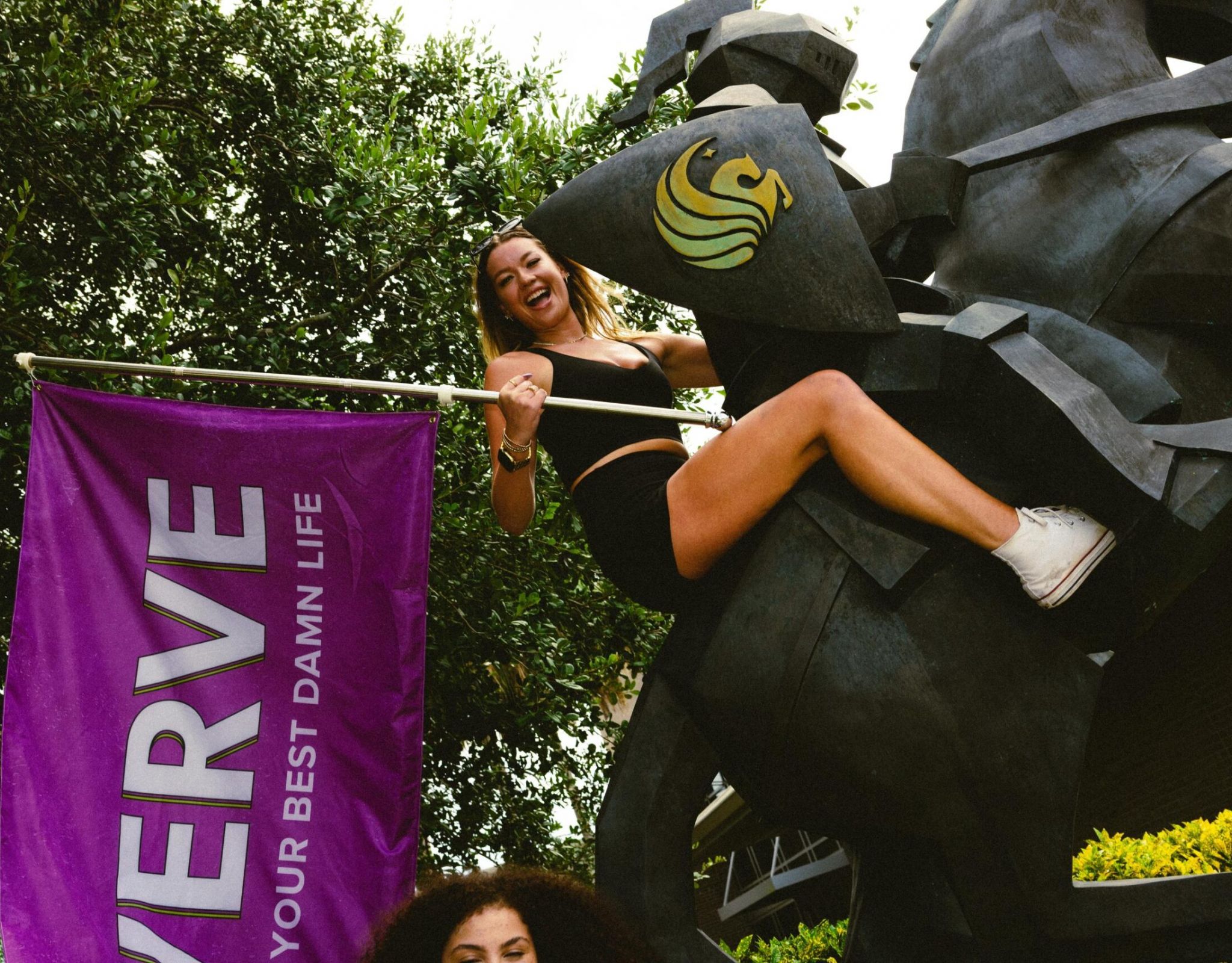 Una mujer de VERVE Orlando, vestida con pantalones cortos negros, posa junto a una estatua de un caballo, junto a una pancarta morada con la inscripción «VERVE», al aire libre.